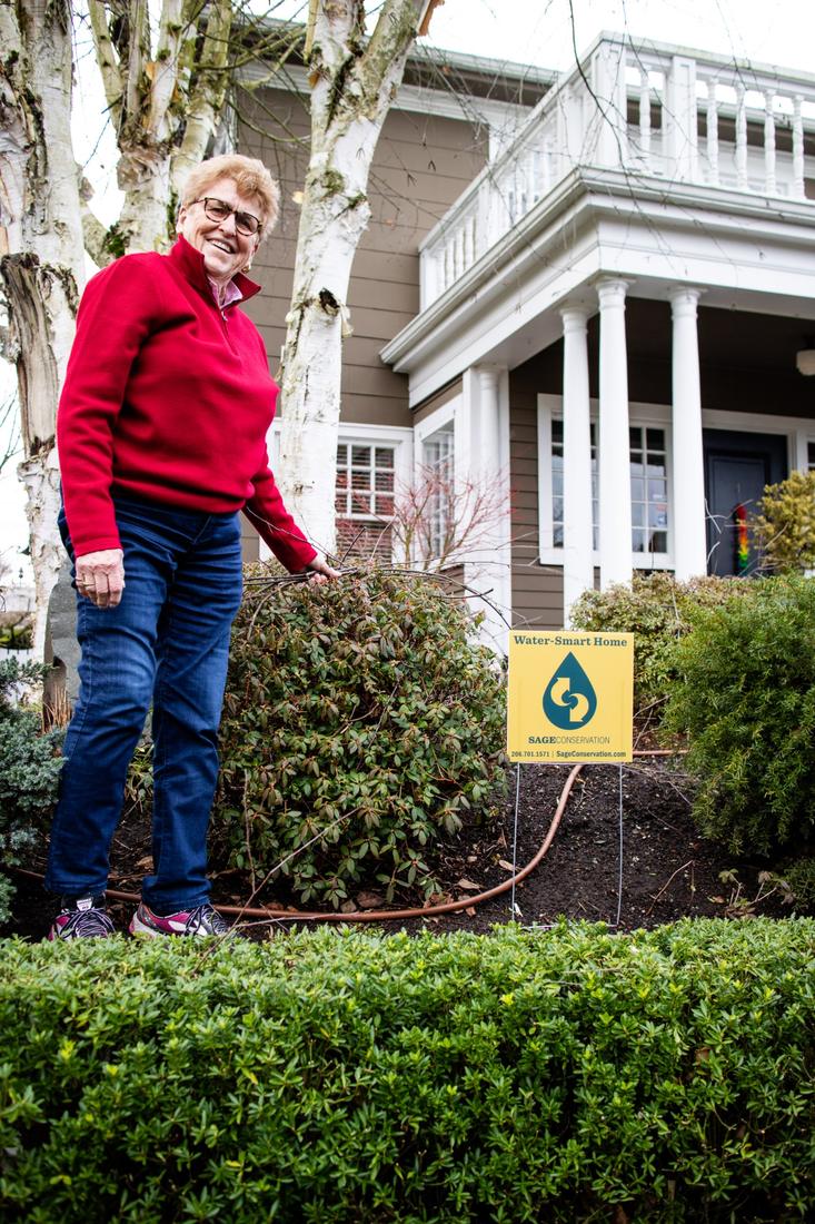 A smiling homeowner in her front yard next to a Water-Smart Home sign